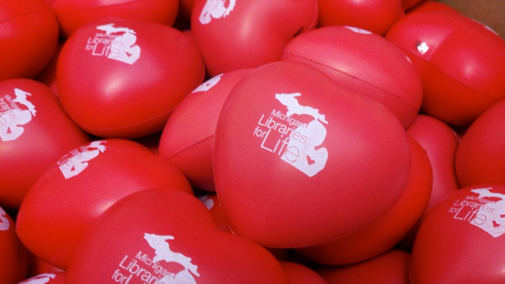 closeup image of a pile of red, heart shaped objects imprinted with an all-white map of the State of Michigan and the words Michigan Libraries for Life