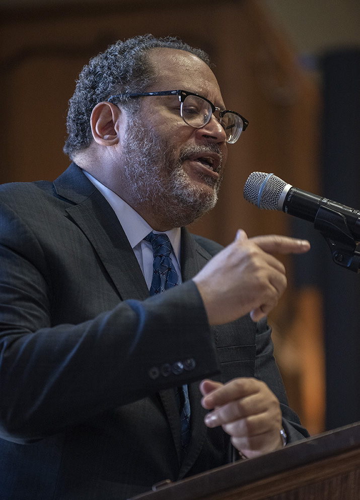 Photo of an African American man from the waist up, speaking into a mic at a lectern