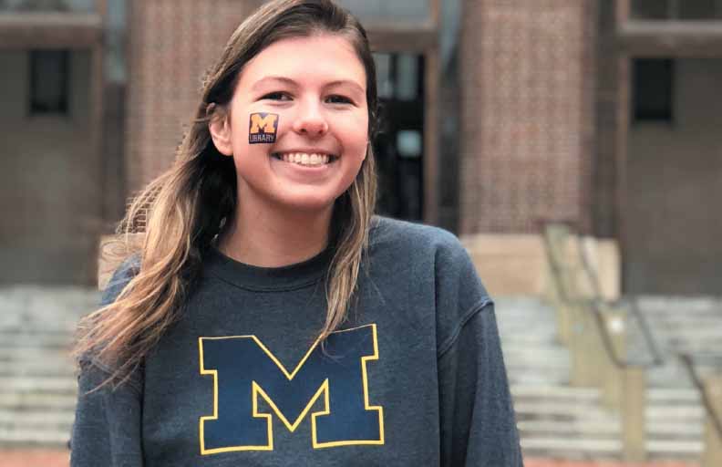 Photo of a woman wearing a tattoo of the library logo on her left cheek and a sweatshirt with a block M on it 