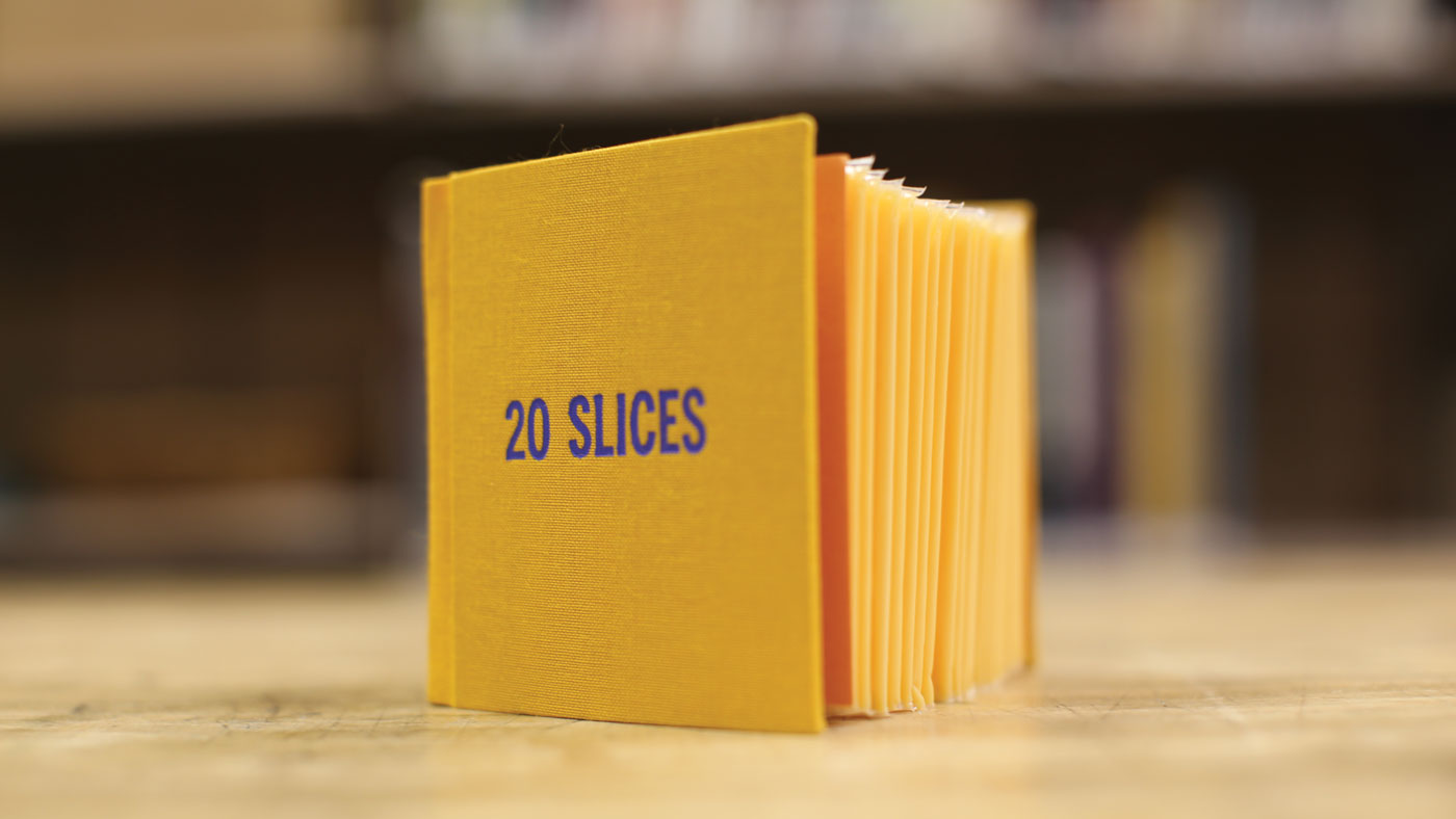 A cloth-bound yellow volume of bound cheese slices, titled 20 Slices, standing upright and open so cheese slices are visible