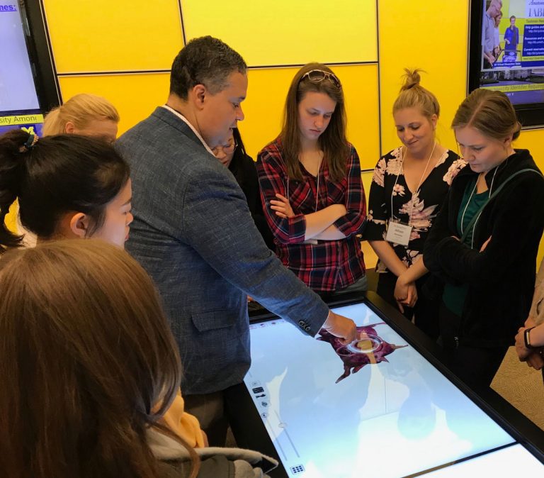 Photograph of a small group of people standing around a horizontal screen, with one person appearing to touch it