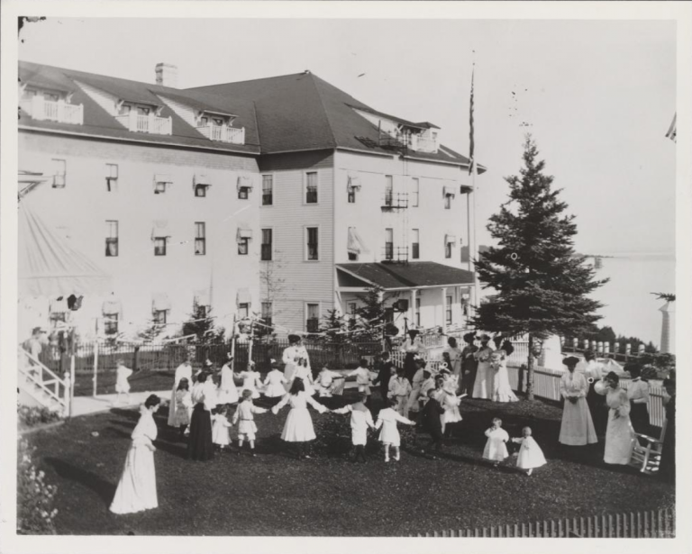 Black and white photograph of a portion of the outside of what appears to be a large white building, and a group of children and adults in the foreground. The women are in long formal dresses suggestive of the early 20th century.