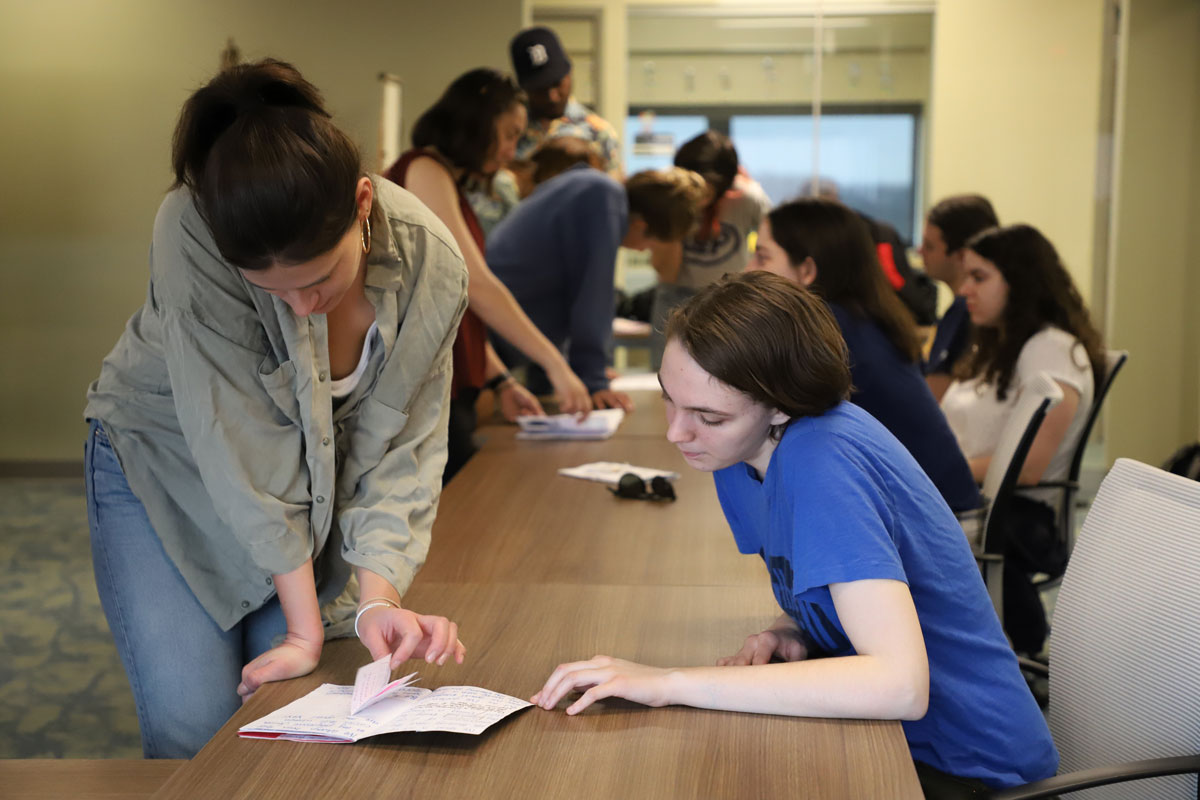 two people together examining an open booklet on a table, with other people visible in the background