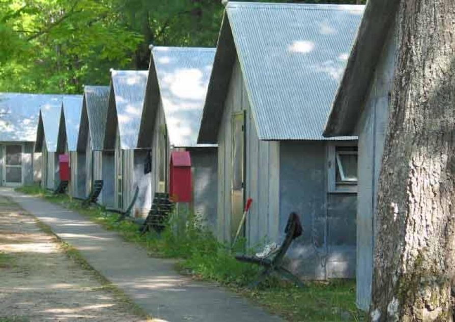 Photo of a row of 8 identical small buildings, with the trunk of a tree in the foreground and a paved path in front of them