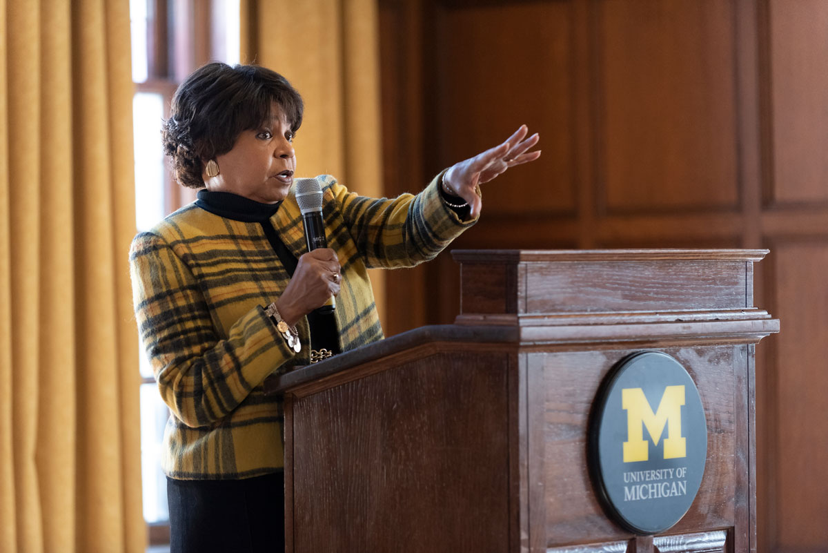 A woman standing at a podium, holding a microphone in one hand and gesturing with the other